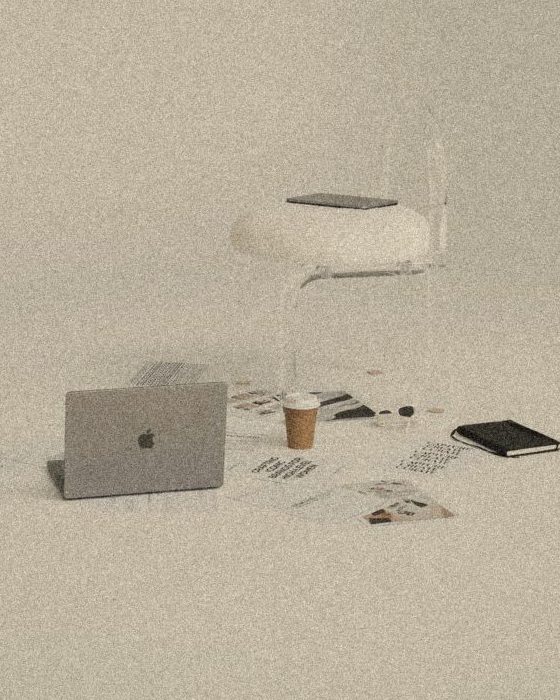 A calm studio scene: a laptop, coffee, notebook and printed pages on the floor beside a sculptural chair
